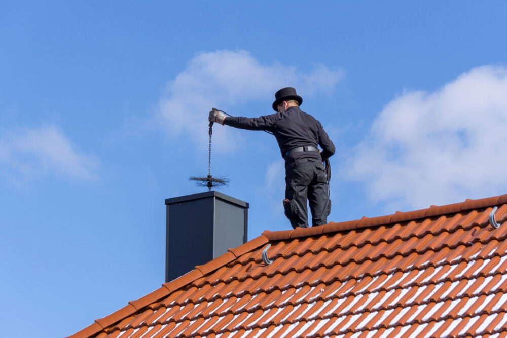 homeguide-chimney-sweep-cleaning-a-chimney-standing-on-the-house-roof-lowering-equipment-down-the-flue
