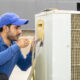 a professional electrician man is fixing a heavy duty unit of central air conditioning system by his tools on the roof top and wearing grey color of uniform and white cap