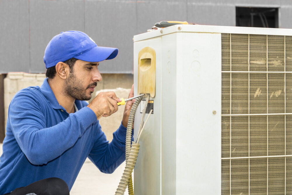 a professional electrician man is fixing a heavy duty unit of central air conditioning system by his tools on the roof top and wearing grey color of uniform and white cap